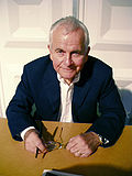 A color photograph of a man in a suit, sitting at a brown desk, holding a pair of glasses