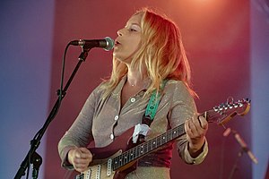 Upper body shot of Jack River, a 27-year-old woman. She is singing into a microphone with her head slightly tilted up and is seen in mostly left profile. She is also playing an electric guitar. The artist is wearing a champagne coloured top, which is sparkling. Her fair-to-blonde hair is over her shoulders. The background has alternate panels of purple-blue and plum sections.