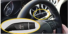 A rectangular image showing the upper part of a black automobile steering wheel, with a hand at lower right and a dashboard visible in the background. A yellow ellipse is around a small lever protruding from the right of the steering column behind the wheel; in the lower left quarter of the image is a closer view of this lever, also within a yellow ellipse. On the lever the letters R, N and D in white are arranged top to bottom in a central column with triangles pointing in opposite directions next to them on the left, and a letter P with a triangle pointing away from it on the right.