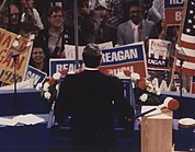 Photograph of Reagan from the back on stage speaking to a large crowd, many holding Reagan signs