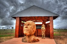 An orange-coloured stone bust of a bolding elderly man looking upwards and smiling with one eye open. The bust fronts a brick pavilion with a triangular grey roof.
