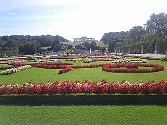Frontale Farbfotografie eines Blumenbeetes mit roten und weißen Blumen, die auf einem Rasen arrangiert sind. Im Hintergrund zwei Fontänen, die vor einem Hügel mit Bäumen und einem Gebäude darauf sind. Rechts sind Hecken mit Skulpturen auf Sockeln
