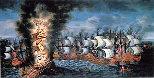 A colorful oil painting showing a large group of sailing warships engaged in battle. In the foreground to the left, a very large ship flying a Swedish flag is listing heavily and a huge explosion is shattering her structure and throwing men and equipment upwards in together with flames and black smoke