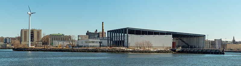 The exterior of the Material Recovery Facility viewed from the other side of the Gowanus Bay. At one side is a large wind turbine, with the administrative building, main processing building, and tipping building on the right