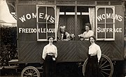 Image of a caravan with white writing on the side reading "Women's Freedom League" and "Women's Suffrage". Two women are seated inside the caravan looking out of an open window, in which two posies of flowers can also be seen. Two other women in long dark skirts and high collared white blouses are stood in front of the caravan.