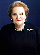 Madeline Albright wearing a dark blouse and coat, with an eagle badge on her left shoulder