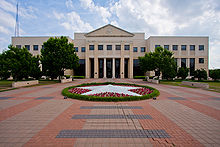 Fronts picture of a two-story administration building on a cloudy day. The walkway is shown leading up to the building including a circular garden in with white flowers forming a star.