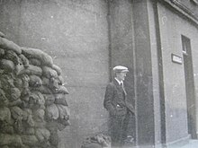 "Black and white photograph of O'Malley wearing a suit and tie and cap, with hands in pockets, looking partly away from the camera as he stands outside a tall building fortified by sandbags to the left"