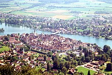 Aerial picture showing a small walled city dotted with red tile roofs and steeples; a wide river runs between it and another city. The farmlands on the far side are bathed in sunlight.