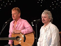 Two grey-haired men sitting at microphones, one holding a guitar