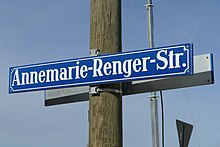 Foto eines blauen Straßenschildes mit weißer Schrift "Annemarie-Renger-Straße", das an einem Holzpfahl befestigt ist