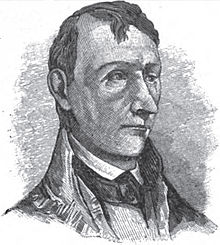 A man with short, dark hair wearing a high-collared white shirt, a high-collared, black jacket, and a black tie