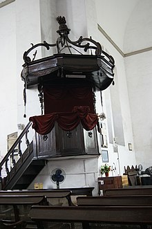 This is an old darkwooden pulpit with red velvet curtains. On the pulpit the minister preaching stands higher than the sitting congregation.