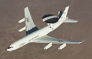 Aerial view port view of white jet aircraft in-flight. It has a large disc-like black radar lying horizontally above two convergent struts.