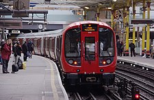 A London Underground S Stock train departs Farringdon with a Metropolitan Line service to Aldgate, with people waiting on the platform to board the next train.