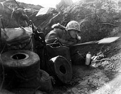 A Caucasian soldier wearing a camouflage helmet and thick winter clothing lies on his side in a trench facing the right of the photo, holding a canteen in his left hand and using an entrenching tool with his right hand. The soldier is surrounded by rolls of communication line, jerries, and boxes of equipment. Behind him, the skyline can be seen above the top of the earthworks.