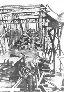 A black and white photograph with a view along a damaged steel girder bridge from one end