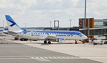 Frontale Farbfotografie von einem blau-weißen Flugzeug mit der Aufschrift „Estonian Air“, das links neben dem Abfertigungsgebäude steht. Rechts stehen Fahrzeuge und hinter dem Flugzeug ist links ein weiteres zu erahnen.