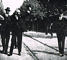 A black-and-white photograph of man wearing a suit, with a bowtie, waving from a railway platform