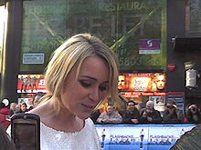 A blond haired woman in a white dress facing right with a large group of people in the background.