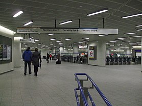 a large white tiled space with a range of ticket barriers in the distance, overhead signs provide directions towards Underground lines