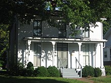 Photo of a white, multi-story house. Partially obscured by bushes and a tree. Four stairs with a handrail on each side lead to the front door.