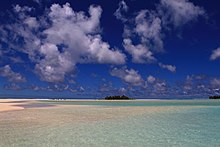 Very shallow water over a white seafloor with a green island in the background beneath blue sky and scattered clouds