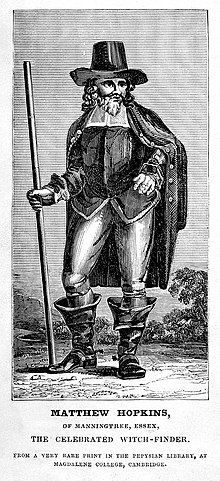 Black and white image of Hopkins. He holds a stick in one hand and has the other placed on his hip, and wears a large hat and wide boots.