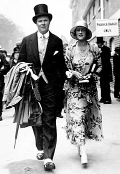 A man in a top hat and suit and a woman in a hat and dress walk towards the camera
