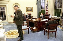 George H. W. Bush sitting at the C&O desk in the Oval Office with Colin Powell on a secondary phone stored in one of the desk's drawers.