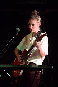 Three-quarter shot of a woman playing an electric guitar in front of her is a microphone on a stand.