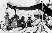 A number of men dressed in shorts and shirts with sleeves rolled up, one in a singlet sit in the shade of an awning. Several wear pith helmets, one of whom sits in the sun shirtless outside the awning leaning against a pole holding up the awning.