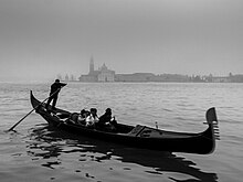 A black-and-white photo of what appears to be a cool, gray day. Four people, indistinct, sit in the middle of a long, thin boat, the gondola, moving to the right. High on the stern (left) stands a man with both hands on a long oar, which disappears into the water to his rear and slightly to his right. In the distant background, across the calm water, the buildings of Venice can be made out, standing in the mist.