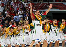 Frontale Farbfotografie einer Mannschaft in weißen Trikots, die sich in einer Reihe in den Armen halten und sich vorbeugen. Vor ihnen streckt ein Spieler seine Hände in die Höhe. Alle haben die Nationalflagge Litauens um ihre Schultern geknotet. Im Hintergrund ist eine Tribüne mit Zuschauern.