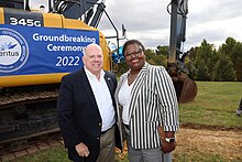 Governor Larry Hogan and Brenda Thiam stand in front of an excavator with the banner "Groundbreaking Ceremony 2022"