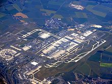 Farbige Luftaufnahme eines Flughafens mit vielen Landebahnen und Terminals. Am oberen Bildrand und unten rechts sind Felder und kleine Orte.