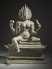 A reddish bronze statue of a four-armed topless woman wearing a short pant sari, standing on a pedestal. She wears a flaming crown and various ornaments. She carries in her hands a goad, a noose, a bowl and a small broken trident.