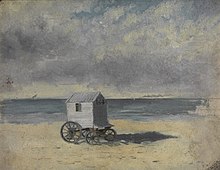 Eine Strandszene unter einem bewölkten grauen Himmel. In der Mitte des Bildes steht ein kleiner Badewagen mit einem Satteldach, einem Fenster und vier Rädern auf dem ansonsten leeren Strand vor einem blaugrauen Meer.