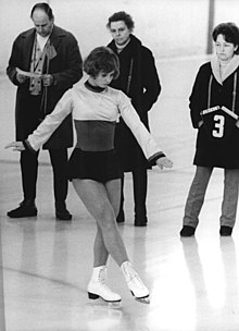 Black and white photo of a young woman, dressed in a short dress and white ice skates, executing figures on ice, while three people, two men and one woman, look on behind her