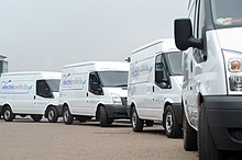 Four white cargo vans, based on the third-generation facelifted Ford Transit but with electric powertrains, made for left-hand traffic, each with the words "electric vehicle" visible in big letters on its right side and in smaller letters on the front-facing part of its raised roof, and with the 2011 logo of the University of Warwick visible on the right (driver's) door