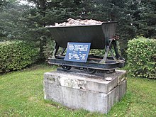 A monument commemorating the beginning of the oil shale industry, consisting of a grey concrete block about 1.5 metres wide and three-quarters of a metre deep. The block is topped by a black metal hopper filled with rocks. A blue plaque with white Estonian-language lettering just above the block explains its significance.