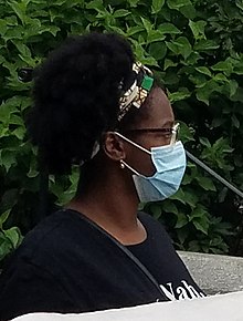 A Black woman with glasses and a white, green, and black hairband, photographed in profile against a bush. She wears a blue surgical mask.