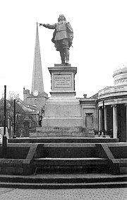 In the foreground is a statue of a man on a plinth above steps, with person sitting on them. In the background is a church tower. The picture is arranged so that the outstretched arm with a pointing finger of the figure appears to be touching the top of the tower.