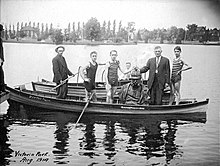 Five men stand in a boat, three of whom are wearing bathing suits. A bust of the Kaiser sits between them. A second boat and man can be seen behind them.