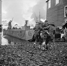 A black and white photograph of a wounded soldier being helped to a landing craft by two other soldiers