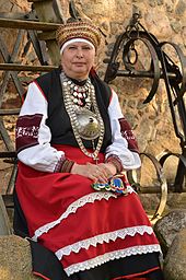 Frontale Farbfotografie von einer älteren Frau in schwarz-weißer Tracht mit rot-weißer Schürze. Sie trägt eine rot-goldene Kopfbedeckung und mehrere Halsketten in Silber. Auf ihrem Schoss befindet sich ein bunt gestricktes Tuch. Im Hintergrund hängen an der Steinwand alte Gerätschaften.