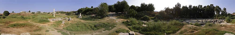 A view of a landscape rising to a hilltop covered with small trees. There are many small hollows, ridges and tracks. The landscape is littered with the remains of marble buildings, including a single column standing to the left.