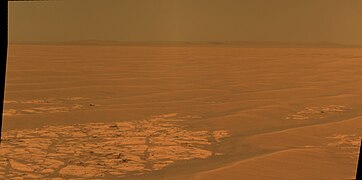 Figure 4. The plain at a field of "plains ripples" soils.[7] Fields of plains ripples cover many square kilometers. The hills on the horizon are the rim of Endeavour Crater.[33] Each plains ripple is a sloped mound of soil, usually around 25 cm tall, 2 m to 4 m wide, and longer than they are wide.[7][34] The ripples' long axes are roughly aligned north-to-south since the prevailing winds are (like the equatorial trade winds on Earth) from east to west. Each ripple can join with others or be separated by patches of lighter Meridiani sediment outcrop (for example, in the left foreground). A scattering of loose blocks sits on the foreground sediment. Most block are ejecta from meteorite impacts. Plains ripples are smaller than dunes, "armored" by a layer of hematite spherules, and, hence, have exceedingly slow migration rates.[7][35][36] Image taken on Sol 2314 (2010-07-28).