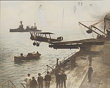 A small biplane has just taken off from a warship, having used a platform built over the roof and barrels of a twin-barrelled turret. Sailors are observing the launch from the deck of the ship, and from a small boat nearby. A large warship is visible in the background.