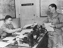 A woman in uniform hands papers to Smith, who wears glasses and is seated behind his desk.